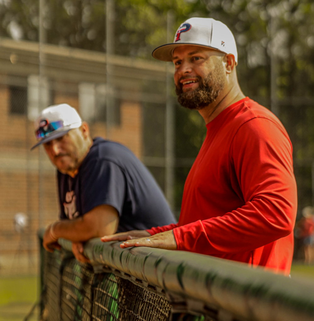 This image captures two USA Prime baseball coaches or team staff standing along the fence, watching the game with focus. The man in the foreground, wearing a red long-sleeve shirt and a white "P" hat, appears engaged, possibly discussing strategy or observing player performance. His facial expression suggests a mix of concentration and encouragement. The second man, slightly blurred in the background, wears a navy blue shirt and a white cap with sunglasses resting on the brim. He is leaning on the railing, watching attentively, adding to the atmosphere of coaching and mentorship. The setting suggests a well-maintained baseball field, with the chain-link fence serving as a barrier between the coaches and the field. The warm lighting and shallow depth of field emphasize the main subject while keeping the background elements contextual.