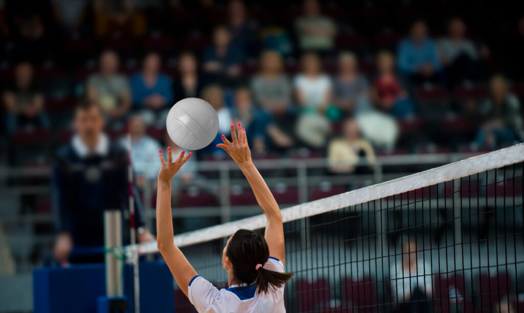 A female volleyball player in a white and blue jersey jumps near the net, setting the ball with both hands. The background features a blurred audience in the stands and a referee watching the play, creating an intense game atmosphere.