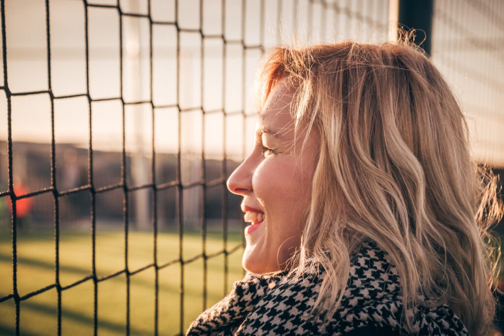 This image captures a blonde woman smiling as she watches something through a sports net, possibly a soccer or lacrosse game. The warm, golden sunlight suggests it’s late afternoon or early evening, casting a soft glow on her face and hair. She is wearing a stylish houndstooth-patterned coat, which adds an element of sophistication. The net in the foreground creates a layered perspective, emphasizing her as a spectator, likely engaged in a game or event happening on the field. The blurred background with green grass and goalposts suggests a sports venue, reinforcing the atmosphere of outdoor athletics.